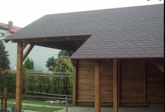Drewniana konstrukcja z dachem pokrytym gontem bitumicznym w kształcie rybiej łuski, widoczna z zewnątrz, z metalowym ogrodzeniem i zielenią w tle.