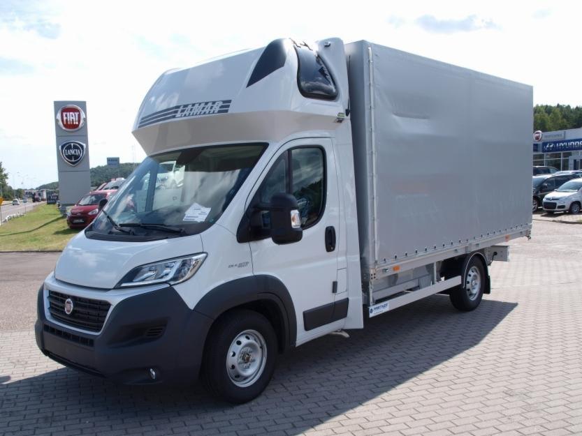 Biały samochód dostawczy z plandeką marki LAMAR na parkingu przed salonem samochodowym Fiat i Hyundai.