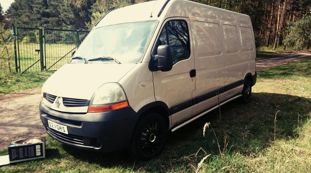 Kremowy van Renault Master z czarnymi felgami zaparkowany na trawie, obok stoi pudełko z elektroniką, w tle ogrodzenie i las.