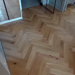 Ułożony parkiet w jodełkę francuską, widoczne przejście do innego pomieszczenia z szarą podłogą i fragmentem białych butów sportowych.