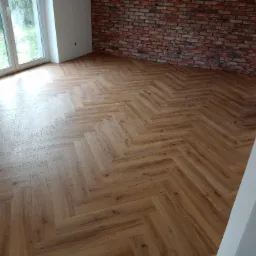 Ułożona w jodełkę podłoga z paneli imitujących drewno w Bytowie, widok na fragment ściany z cegły i okno.