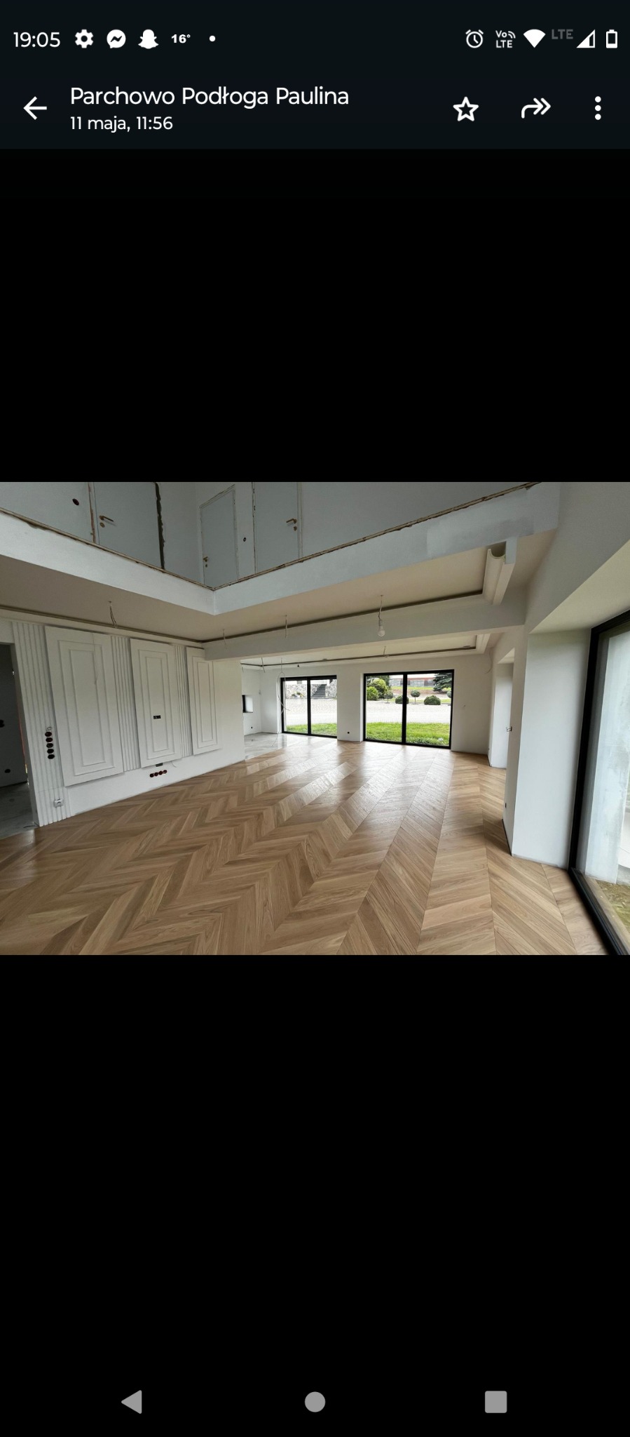 Wnętrze przestronnego salonu z parkietem ułożonym we wzór jodełki francuskiej, białe ściany z dekoracyjnymi panelami, duże okna z widokiem na ogród, sufit z elementami oświetleniowymi w trakcie...