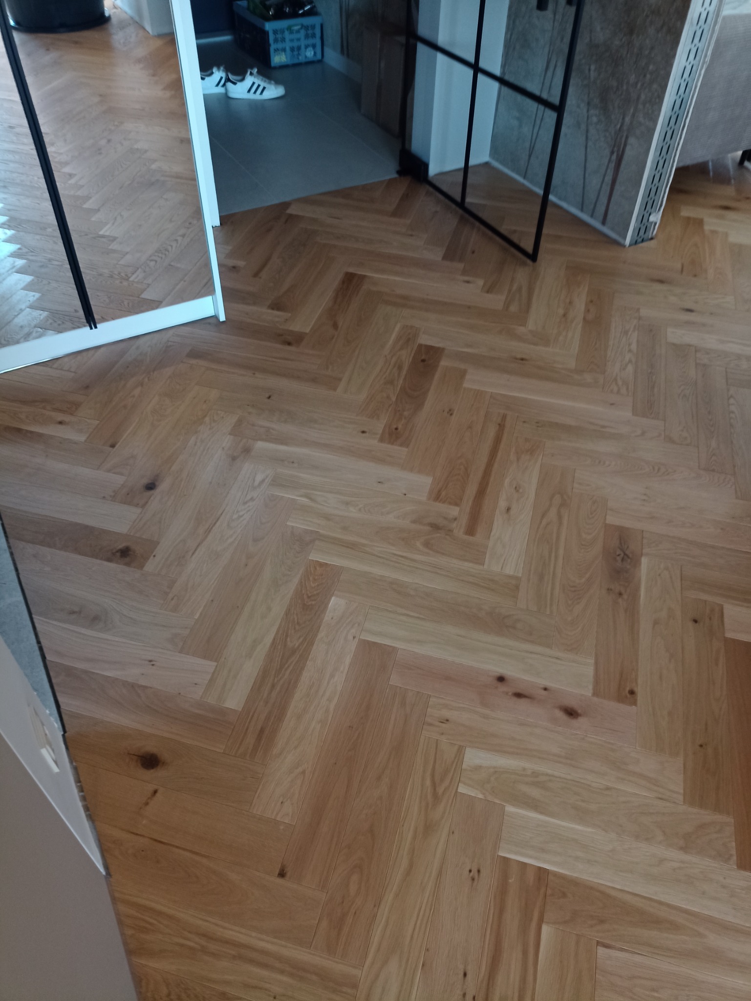 Ułożony parkiet w jodełkę francuską, widoczne przejście do innego pomieszczenia z szarą podłogą i fragmentem białych butów sportowych.