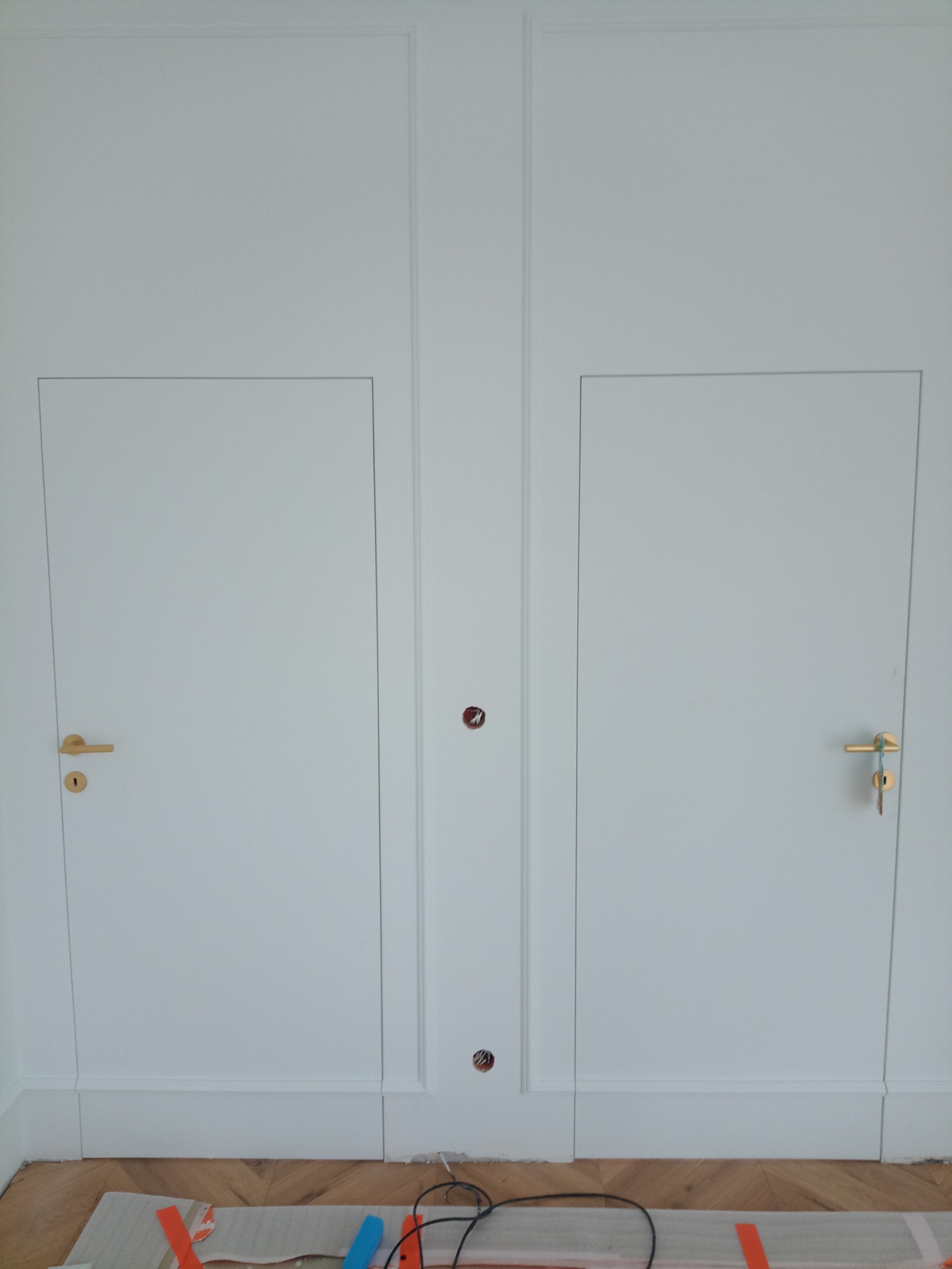 Dwie białe, ukryte drzwi z złotymi klamkami, wbudowane w ścianę z dekoracyjnymi listwami, na podłodze parkiet i kable elektryczne.