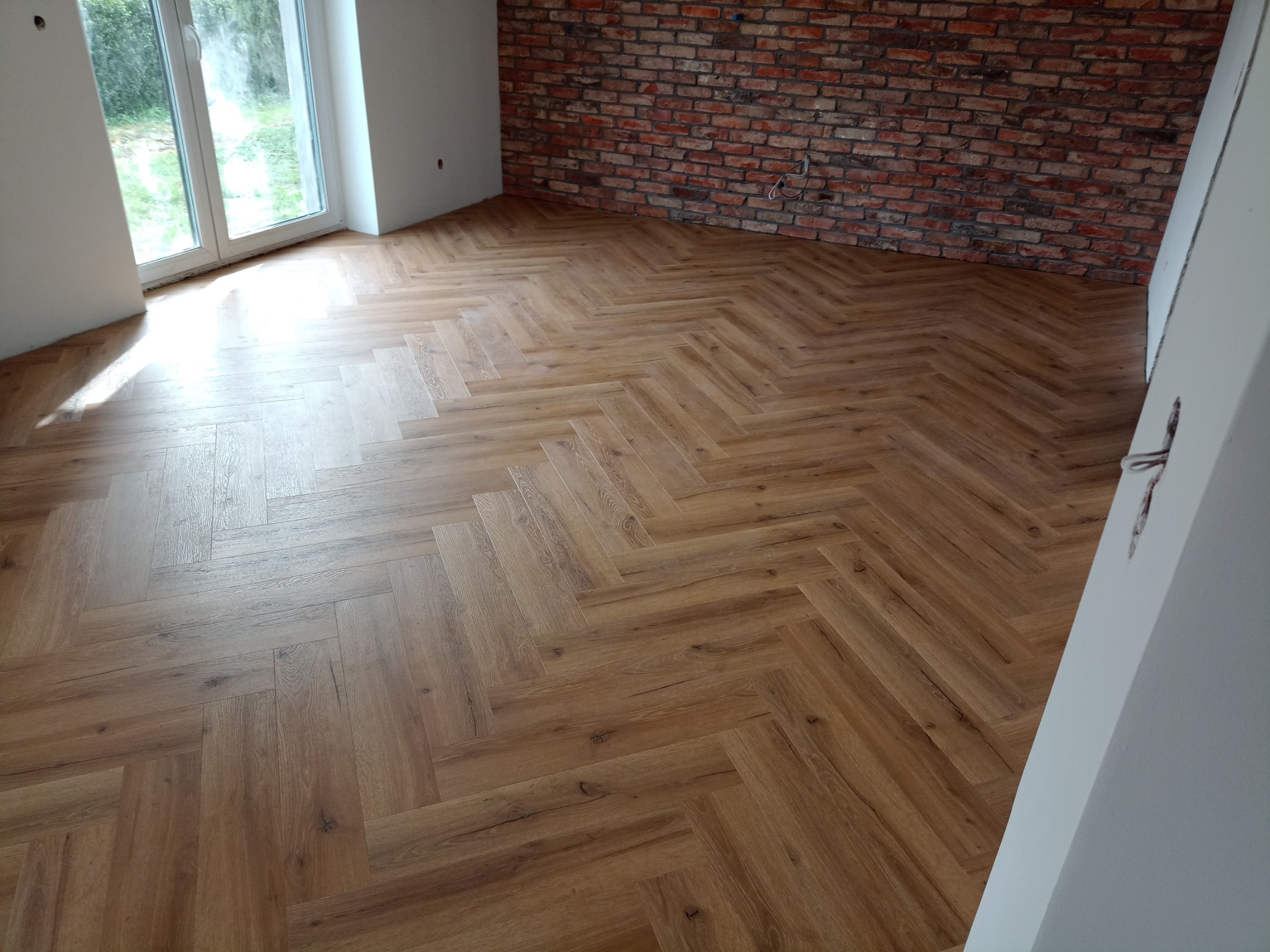 Ułożona w jodełkę podłoga z paneli imitujących drewno w Bytowie, widok na fragment ściany z cegły i okno.