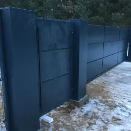 Nowoczesne, grafitowe ogrodzenie panelowe z przesuwną bramą, widoczne na tle zimowego krajobrazu z drzewami iglastymi i częściowo ośnieżonym terenem.
