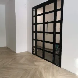 Czarna, metalowa konstrukcja przeszklonych drzwi w stylu industrialnym, z widocznymi spawami i uchwytami, zamontowana w białym pomieszczeniu z podłogą ułożoną w jodełkę.