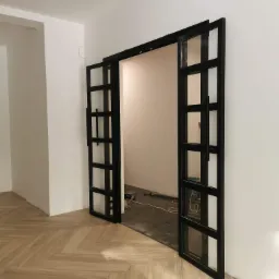 Nowoczesne, przesuwne drzwi szklane w czarnej metalowej ramie, zamontowane w jasnym wnętrzu z parkietem ułożonym w jodełkę, widoczne przewody i narzędzia sugerują trwający montaż.