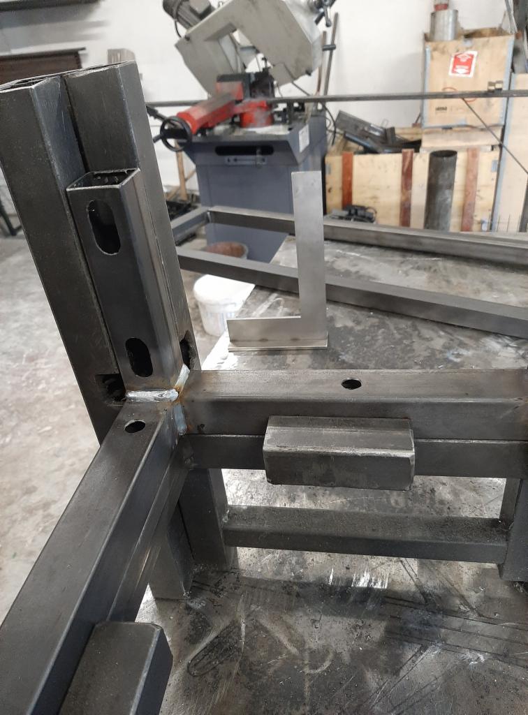 Metalowa konstrukcja spawana z profili, widoczne spawy, kątownik pomiarowy na stole warsztatowym, w tle piła taśmowa do metalu.