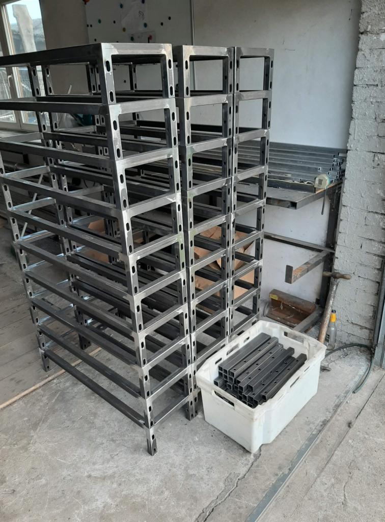 Stalowe regały warsztatowe z profili zamkniętych, ustawione jeden na drugim, obok biały pojemnik z elementami konstrukcyjnymi.