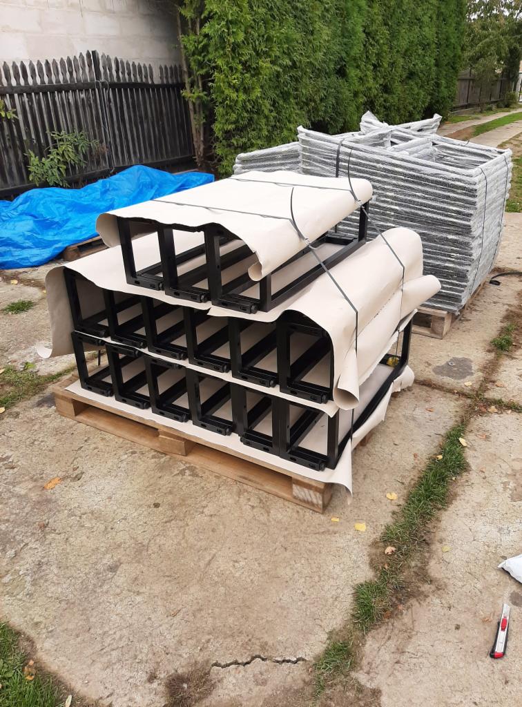 Stosy czarnych, metalowych konstrukcji ramowych na paletach, częściowo owinięte papierem ochronnym i spięte taśmą, ułożone na zewnątrz na betonowym podłożu, z widocznym nożykiem introligatorskim...