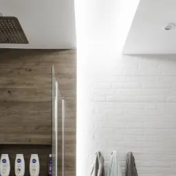 Nowoczesna łazienka z prysznicem typu walk-in, drewnopodobnymi płytkami na ścianie prysznica, półkami wnękowymi z kosmetykami, białą cegłą na pozostałych ścianach, drewnianymi półkami dekoracyjnymi...