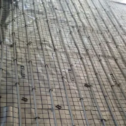 Szare rury ogrzewania podłogowego marki Keller, przymocowane do folii aluminiowej z nadrukowaną siatką, widok z góry podczas instalacji.