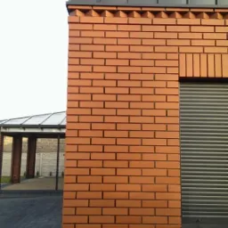 Kamera monitoringu zamontowana na ceglanym budynku z szarym dachem i roletami okiennymi, widok z zewnątrz.