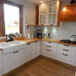 Jasna kuchnia z białymi szafkami i drewnianym blatem, ceramiczny zlew farmerski pod oknem, miedziany okap nad płytą gazową.