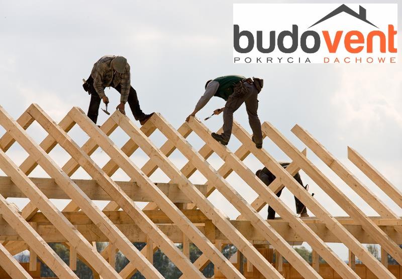 Konstrukcja drewniana więźby dachowej w trakcie montażu, dwóch dekarzy wbija gwoździe, w tle logo firmy budowlanej.