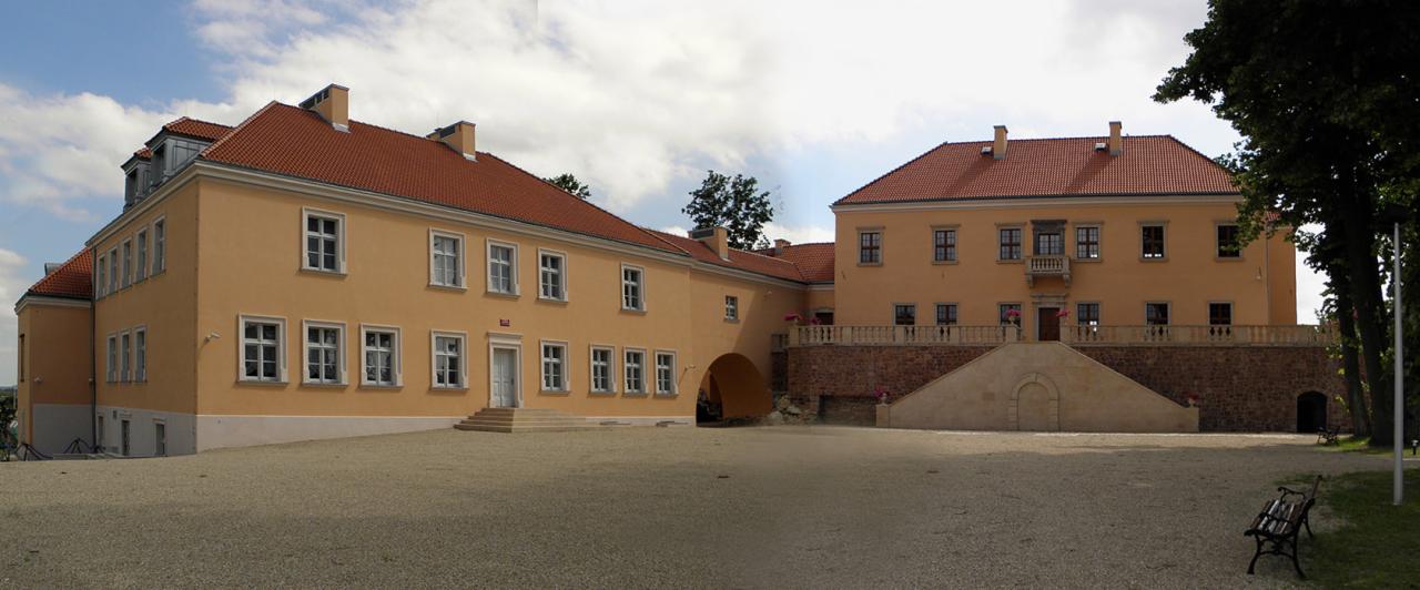 Odrestaurowany, dwupiętrowy budynek z żółtą elewacją i czerwoną dachówką, widoczny z dziedzińca pokrytego żwirem.