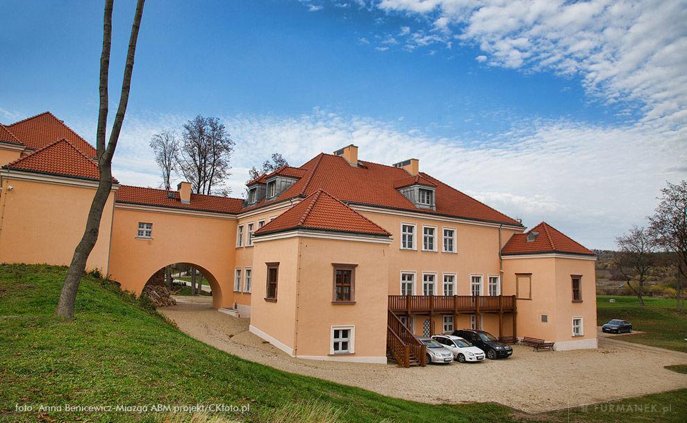 Rozległy, dwupiętrowy budynek z pomarańczową elewacją i czerwoną dachówką, widoczny z poziomu gruntu, z kilkoma samochodami zaparkowanymi przed wejściem i fragmentem zielonego trawnika oraz drzew...