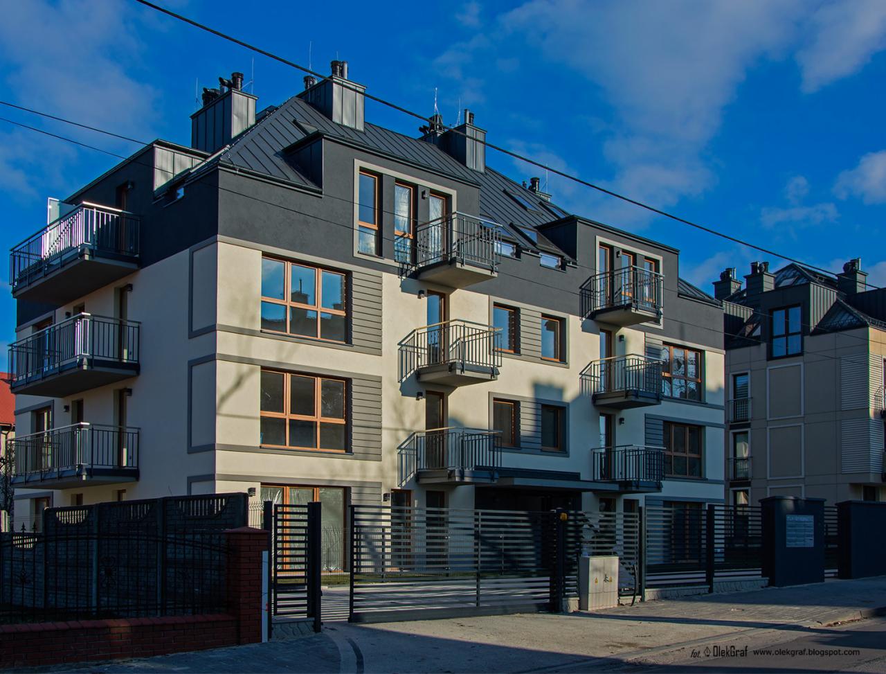Nowoczesny budynek wielorodzinny z balkonami i grafitowym dachem z blachy na rąbek stojący, widoczny z poziomu ulicy przez metalowe ogrodzenie.