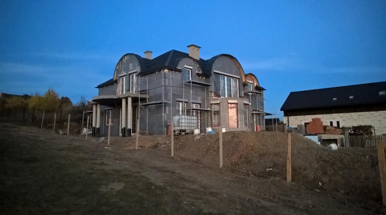 Dwupiętrowy dom w trakcie ocieplania styropianem, z widocznymi rusztowaniami i nowym dachem, sfotografowany o zmierzchu.