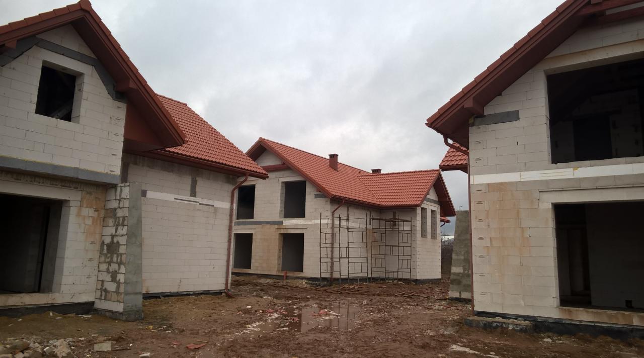 Trzy domy w stanie surowym otwartym z czerwoną dachówką, widoczne rusztowania i błotniste podłoże w pochmurny dzień.