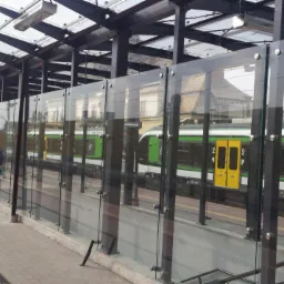 Peron kolejowy z przeszkloną wiatą i barierkami, widoczny pociąg w barwach zieleni i żółci, perspektywa z poziomu peronu.