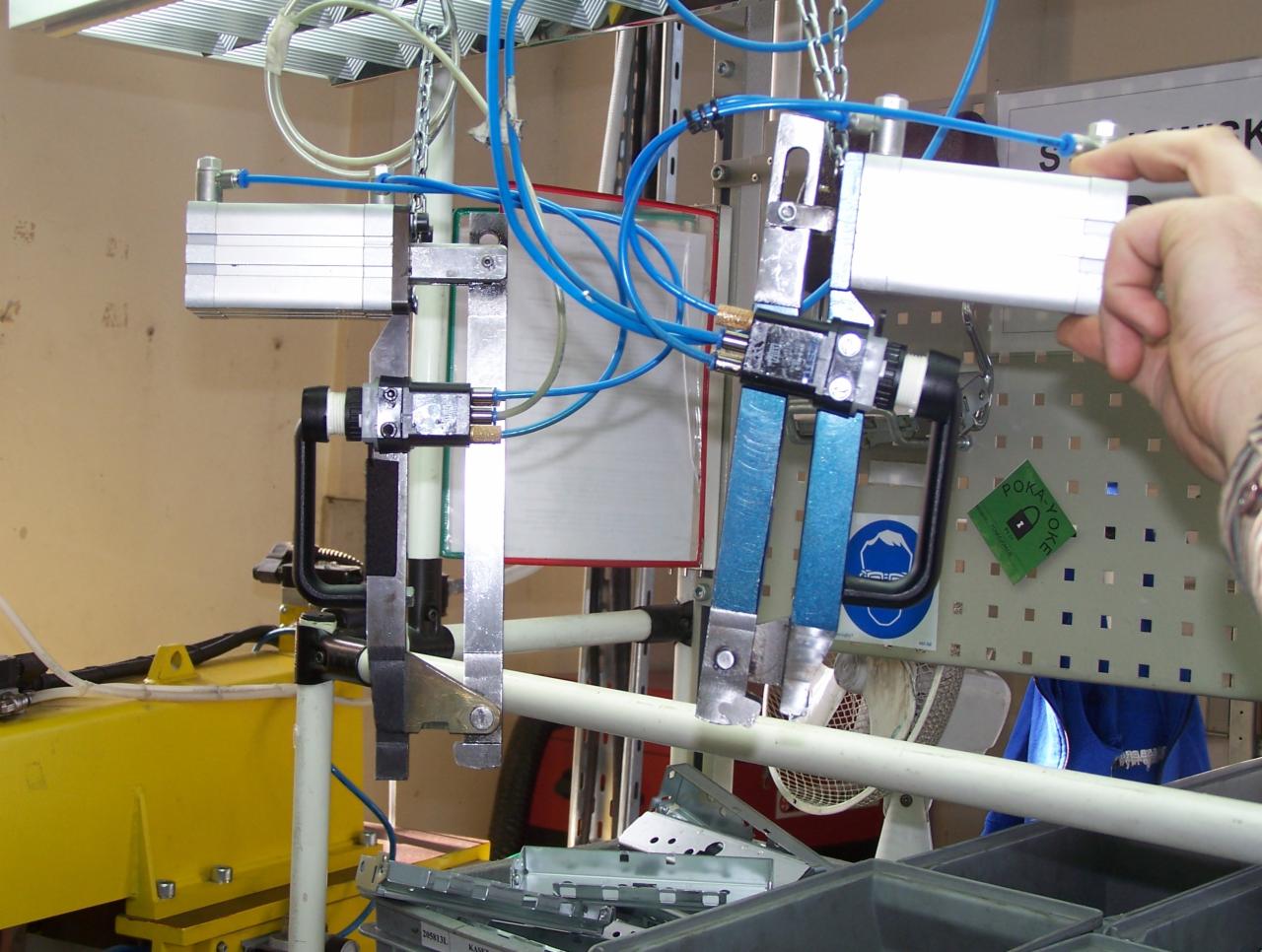 Detale automatycznej linii produkcyjnej z pneumatycznymi siłownikami, widoczne węże i elementy mocujące, w tle fragment hali produkcyjnej i ręka operatora