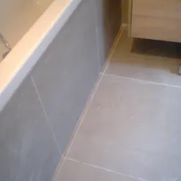 Fragment wykończonej łazienki: szare płytki na ścianie przy wannie i na podłodze, widoczny fragment szafki pod umywalkę z jasnego drewna.