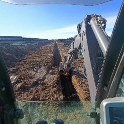 Widok z kabiny koparki podczas wykonywania głębokiego wykopu w ziemi, w tle panele słoneczne i turbiny wiatrowe.