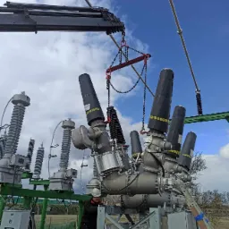 Montaż transformatora wysokiego napięcia za pomocą dźwigu, widoczny pracownik w kasku na drabinie, błękitne niebo z chmurami w tle.