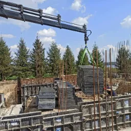 Żuraw przenosi szalunki na placu budowy fundamentów, widoczne zbrojenia, ziemia i drzewa w tle.