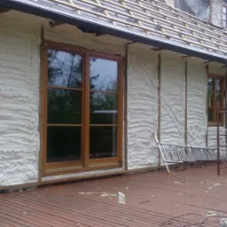 Ocieplanie pianką PUR na ścianie zewnętrznej domu z drewnianą stolarką okienną i widocznym fragmentem dachu przed położeniem dachówki.