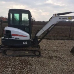 Niewielka, biało-czarna koparka Bobcat E32 na żwirowym podłożu, z widocznym lemieszem i ramieniem, na tle zaoranego pola i zabudowań.