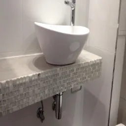 Biała, owalna umywalka nablatowa z chromowaną baterią, zamontowana na blacie wykończonym mozaiką z drobnych, szklanych płytek w odcieniach szarości i bieli, widoczne chromowane rury odpływowe.