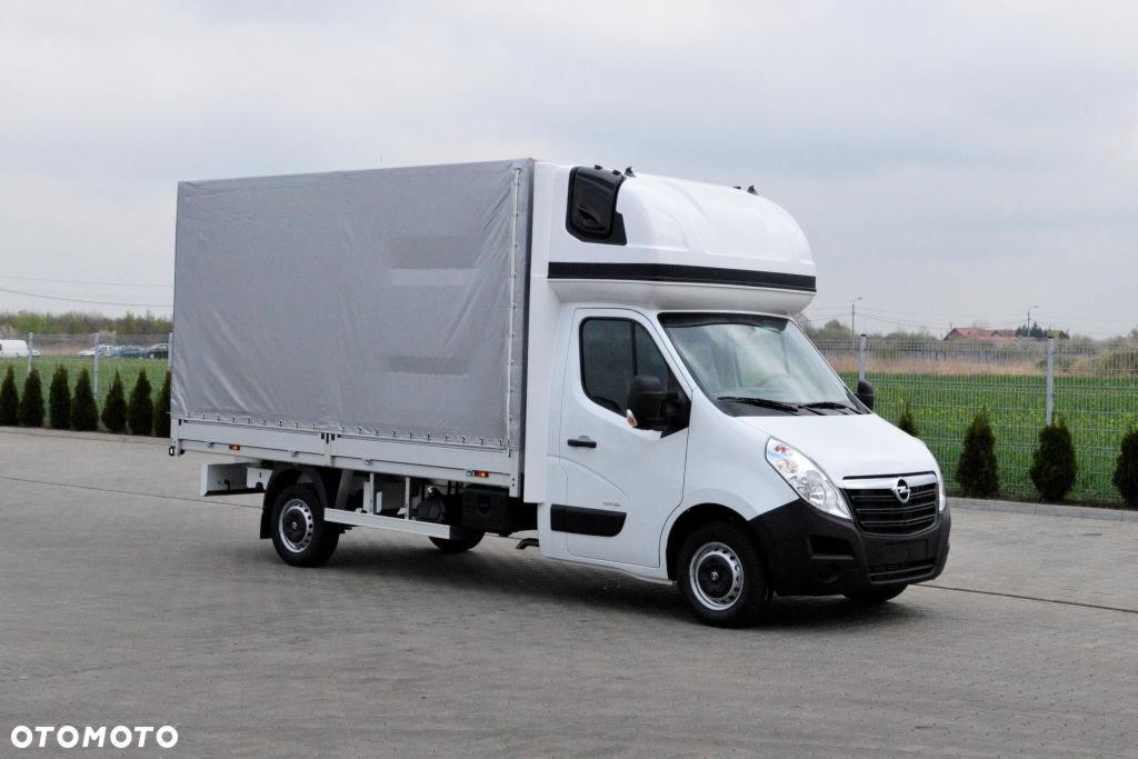 Biały samochód dostawczy z plandeką, zaparkowany na betonowym placu, widok z boku, w tle zieleń i ogrodzenie.