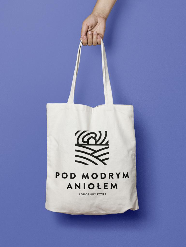 Biała torba bawełniana z czarnym logo przedstawiającym stylizowany krajobraz i napis 'Pod Modrym Aniołem AGROTURYSTYKA', trzymana przez dłoń na niebieskim tle.
