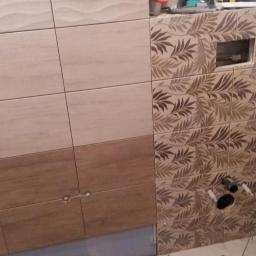 Usługi Remontowo-Budowlane - Ściana łazienki z częściowo ułożonymi płytkami w dwóch wzorach: gładkie beżowe oraz z motywem liści, widoczne przyłącza kanalizacyjne i otwór na instalację.