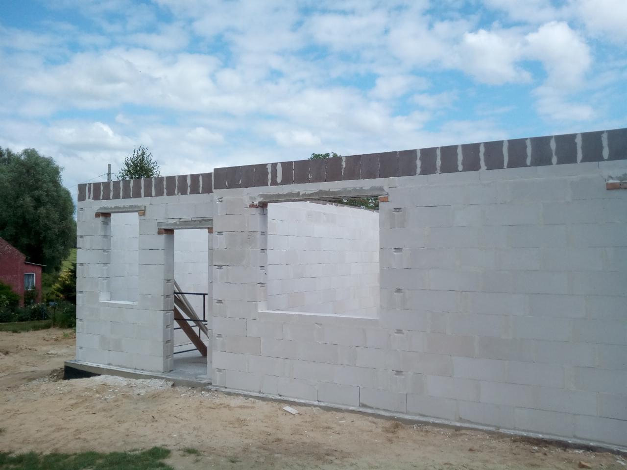 Wznoszone ściany zewnętrzne budynku z białych bloczków, widoczne otwory okienne i drzwiowe, nadproża z ciemniejszych bloczków, teren budowy z piaskiem i trawą, w tle drzewa i fragment czerwonego...