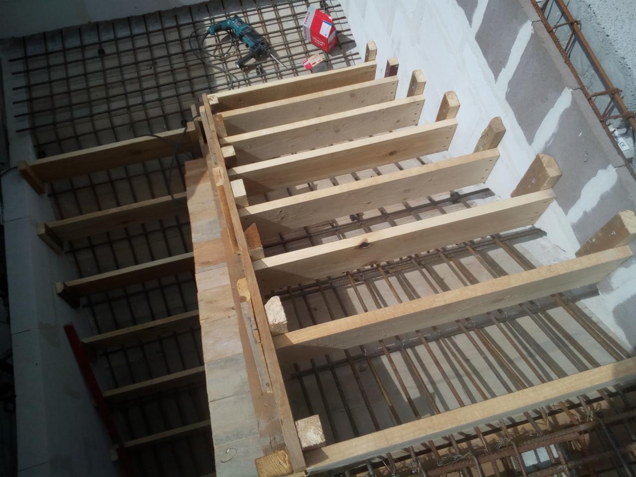 Wewnętrzna konstrukcja schodów żelbetowych w trakcie budowy, widoczne drewniane szalunki i zbrojenie stalowe, w tle wiertarka i puszka z zaprawą.
