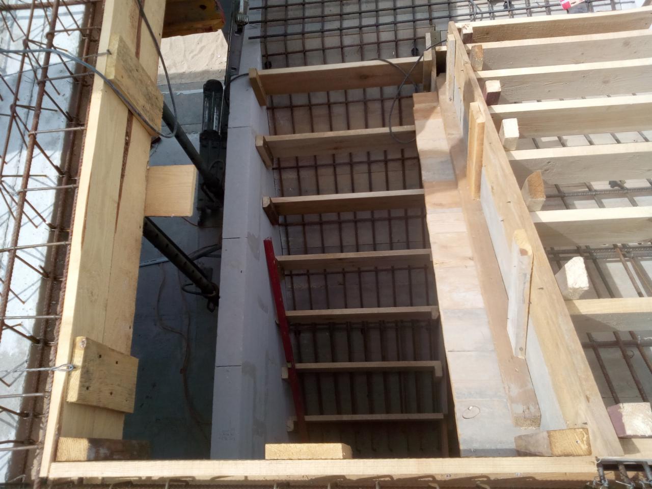 Szczegółowe ujęcie z góry na szalunek schodów z drewnianych desek i stalowych prętów zbrojeniowych, czerwona poziomica oparta o ścianę.