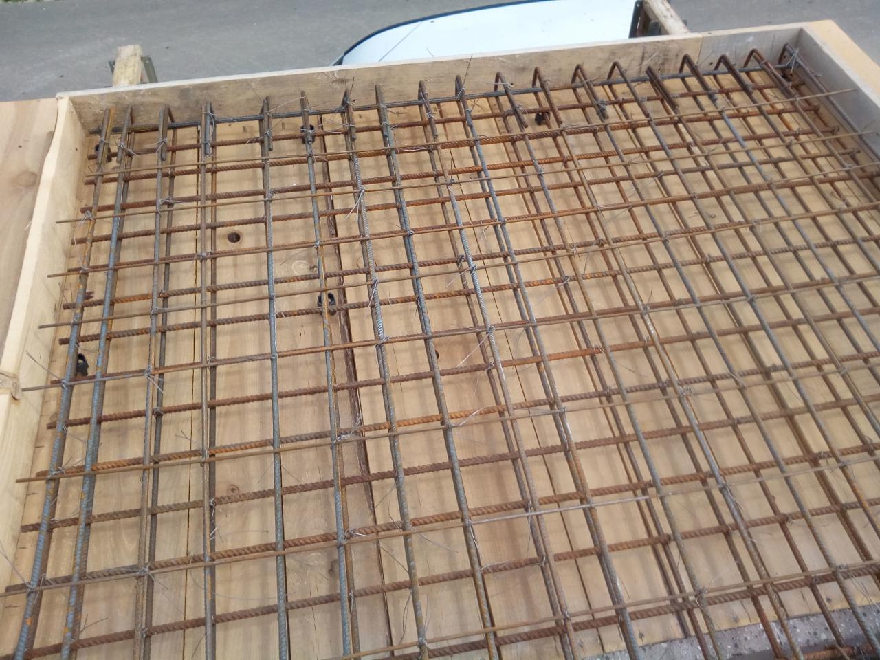 Zbrojenie z prętów stalowych ułożone w drewnianym szalunku na desce, przygotowane do zalania betonem.