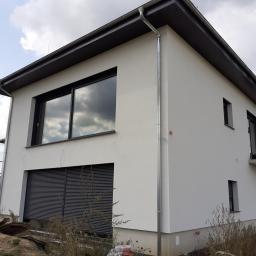 Michał Kałos - Nowoczesny dom jednorodzinny z białymi ścianami, szarymi oknami i roletami zewnętrznymi, widoczne rynny i fragment rusztowania, niezagospodarowany teren wokół.