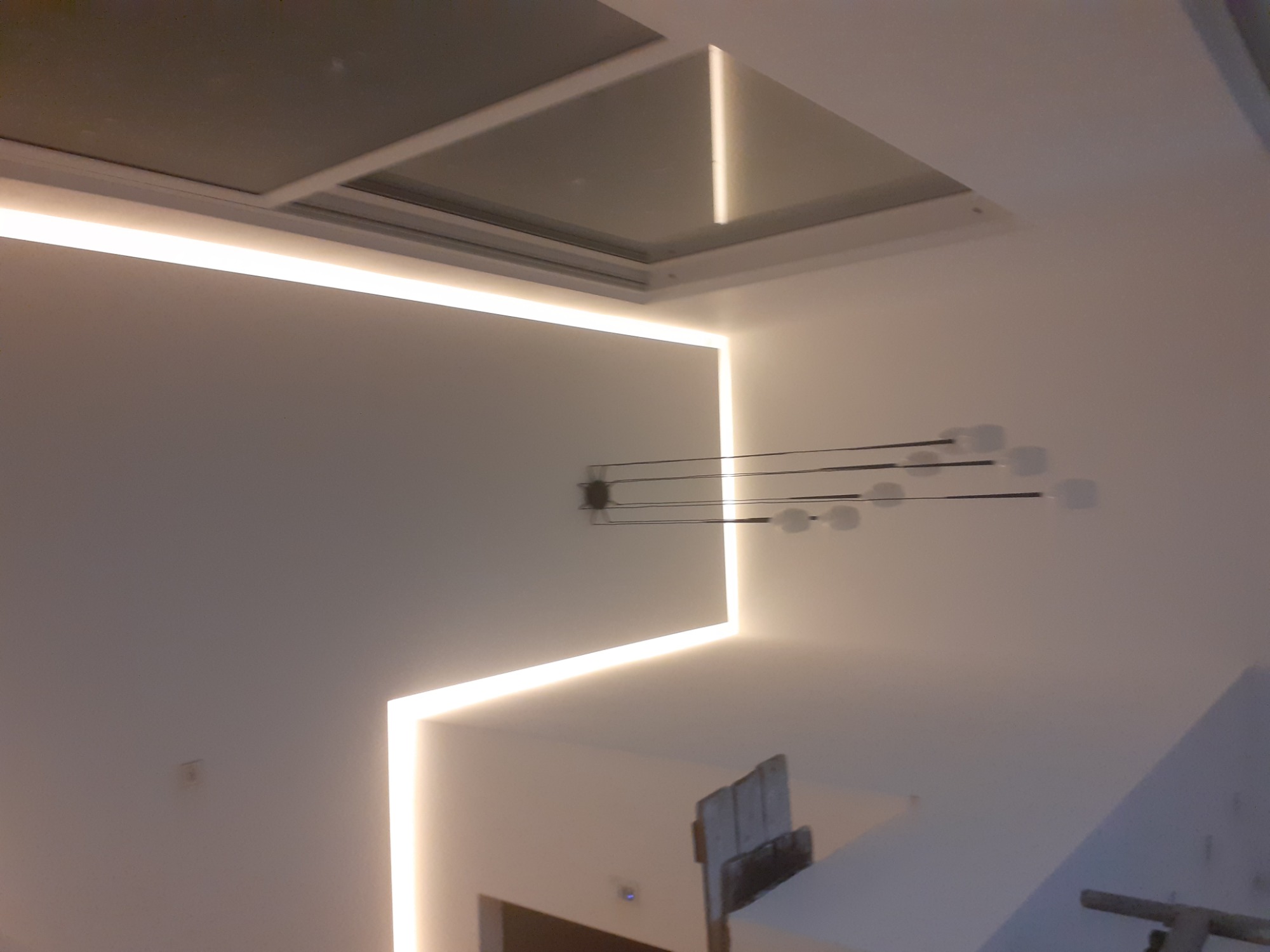 Nowoczesne oświetlenie sufitowe LED w kształcie litery L, z dodatkowym oknem dachowym i minimalistyczną lampą wiszącą z kilkoma punktami świetlnymi.