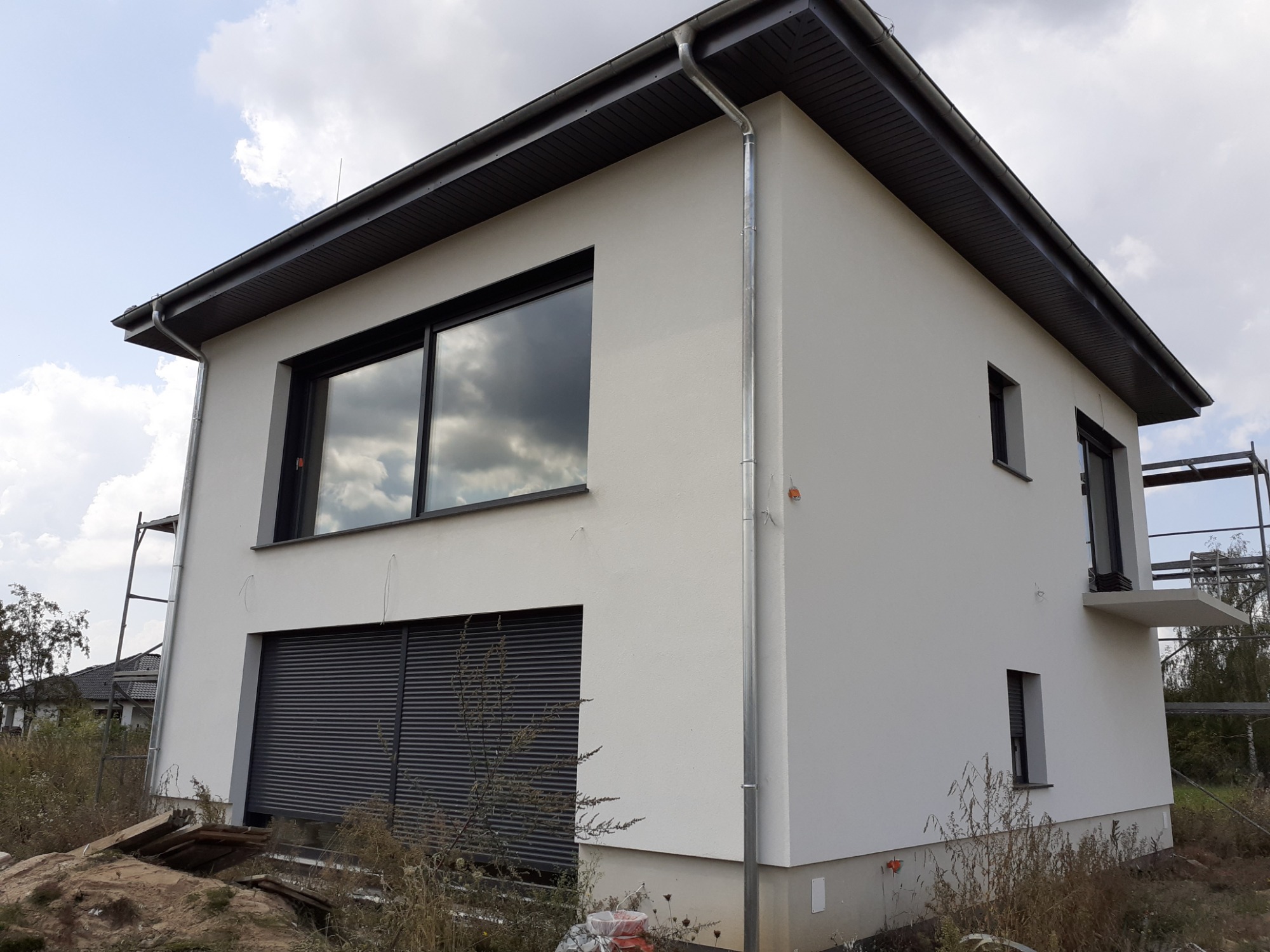 Nowoczesny dom jednorodzinny z białymi ścianami, szarymi oknami i roletami zewnętrznymi, widoczne rynny i fragment rusztowania, niezagospodarowany teren wokół.