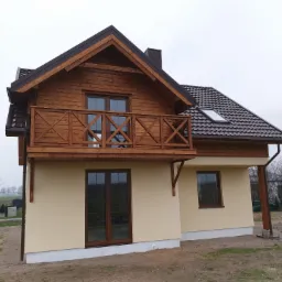 Dwukondygnacyjny dom z drewnianą elewacją na poddaszu, balkonem z drewnianą balustradą i brązowym dachem z dachówki, w trakcie wykańczania terenu.