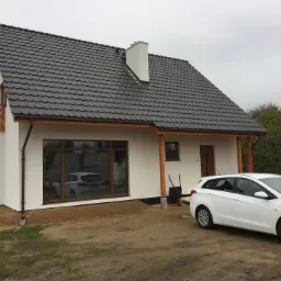 Nowoczesny, parterowy dom z ciemną dachówką, drewnianymi elementami wykończeniowymi i białym samochodem zaparkowanym przed budynkiem na nieutwardzonym terenie.