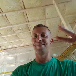 Uśmiechnięty mężczyzna w zielonej koszulce z logo firmy budowlanej, robiący selfie na tle konstrukcji dachu z izolacją z wełny mineralnej i folią paroizolacyjną, trzymający rozłożoną miarkę stolarską.