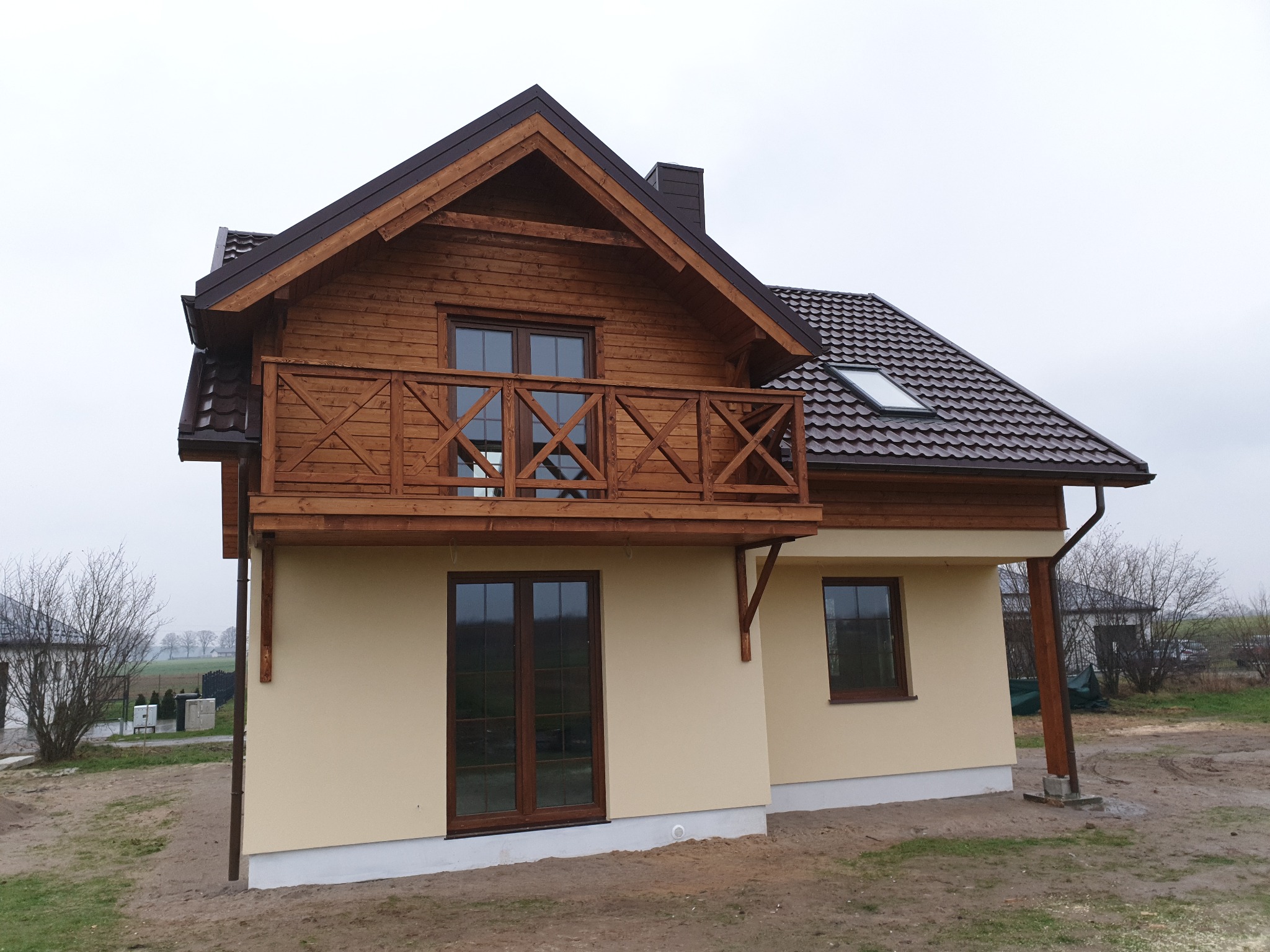 Dwukondygnacyjny dom z drewnianą elewacją na poddaszu, balkonem z drewnianą balustradą i brązowym dachem z dachówki, w trakcie wykańczania terenu.