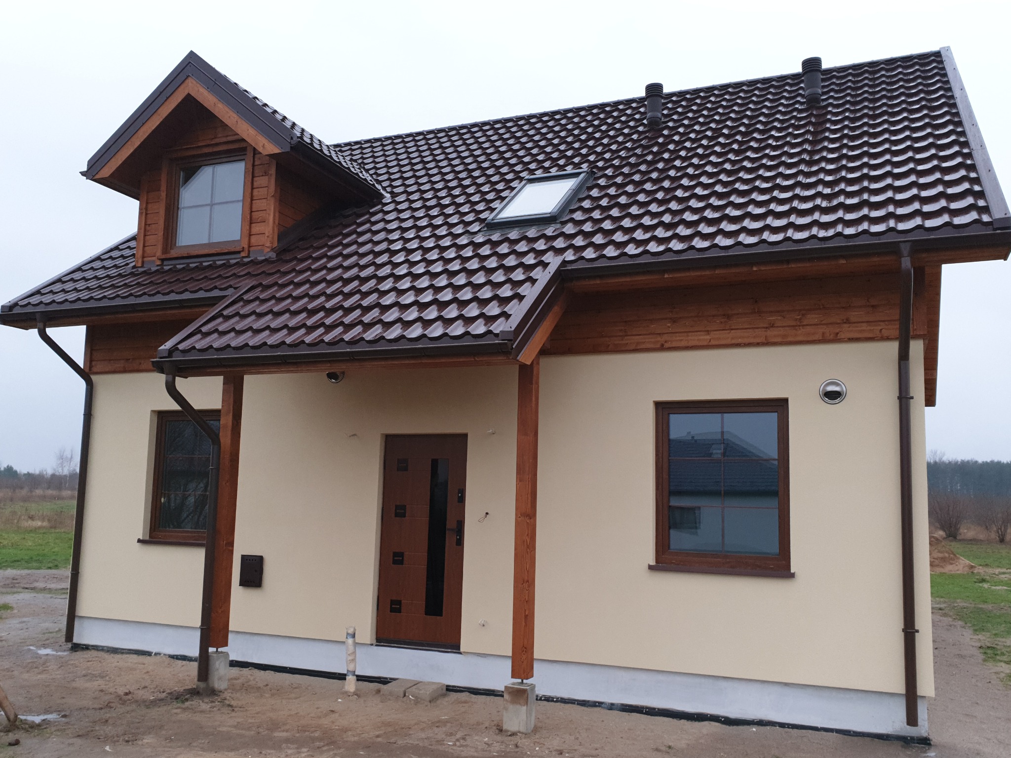 Gotowy dom szkieletowy z brązową dachówką, drewnianą stolarką okienną i drewnianymi słupami podtrzymującymi zadaszenie nad wejściem, widoczny lukarn i okno dachowe.