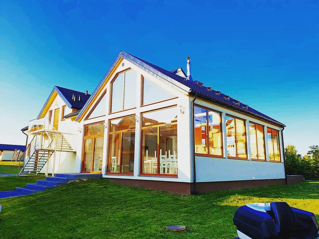 Nowoczesny, parterowy budynek z dużymi przeszkleniami i drewnianą stolarką okienną, otoczony zielonym trawnikiem i błękitnym niebem.